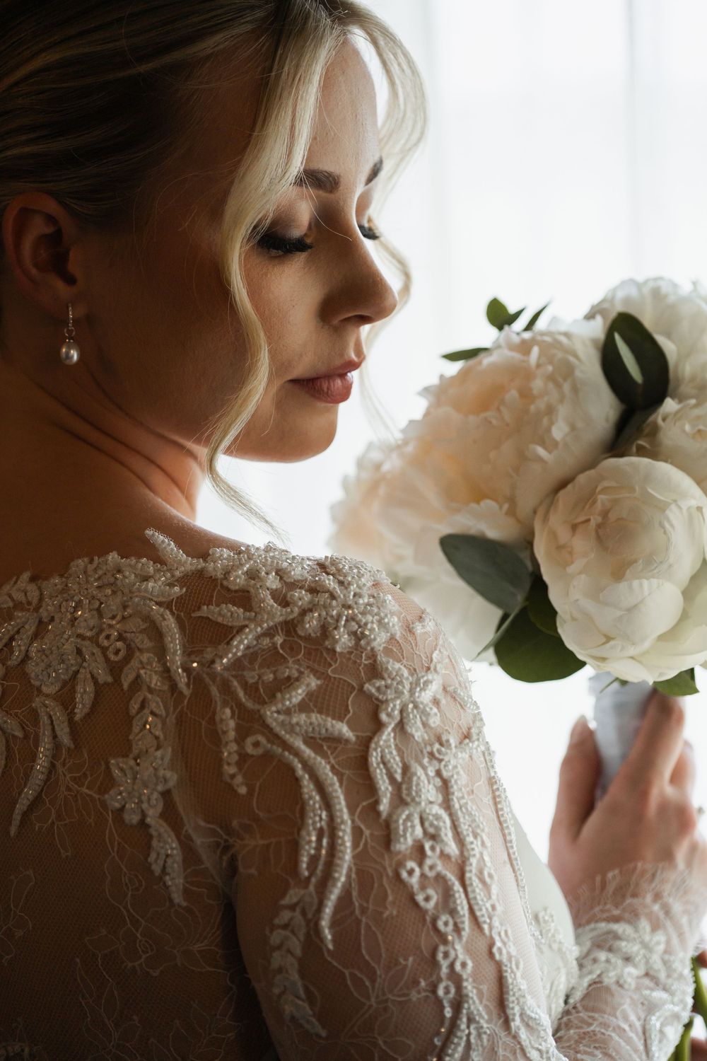 Close up profile view of bride in delicate lace wedding dress holding white peonies against soft natural light.