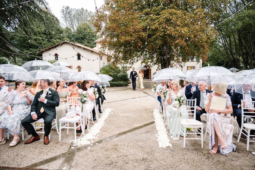 Outdoor wedding ceremony with white umbrellas and guests seated in white chairs along a path leading to a rustic building.
