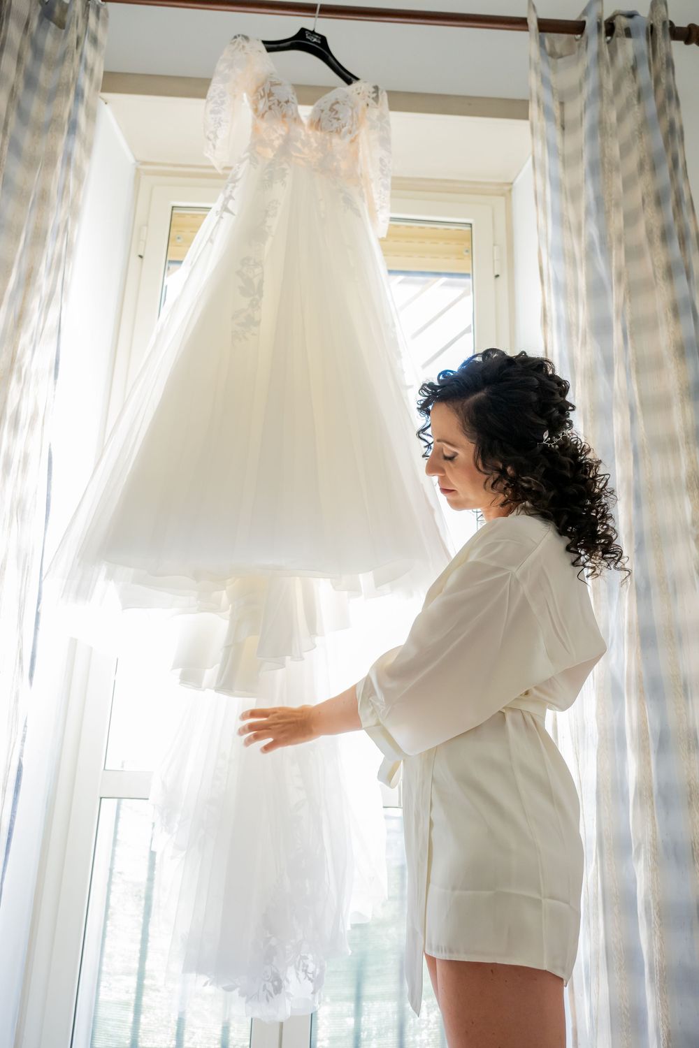 Woman in white outfit admiring wedding dresses hanging on display in front of window with sheer curtains.