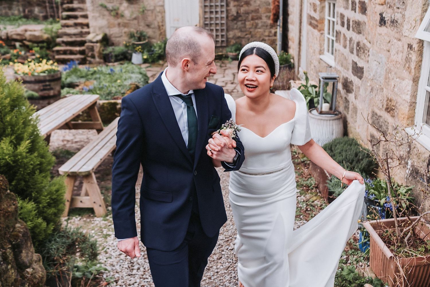 Bride in fitted white dress shares a joyful moment with groom in navy suit against stone wall.