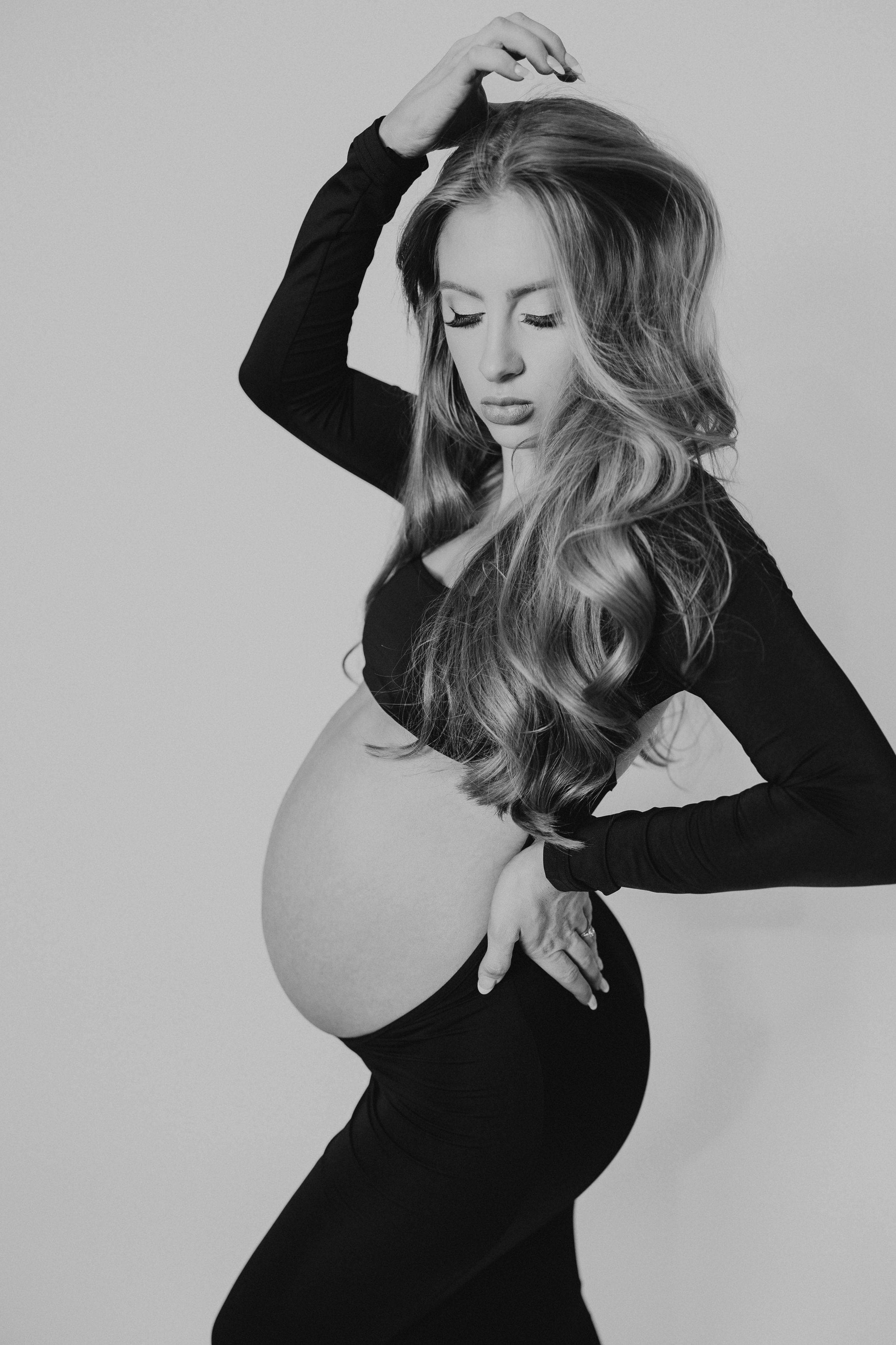 Elegant pregnant woman in black outfit cradling baby bump, black and white studio maternity portrait.