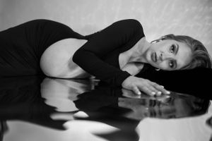 Pregnant woman in black dress lying on reflective floor, artistic black and white maternity photo.