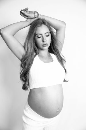 Pregnant woman in white crop top with arms raised, showcasing baby bump in black and white studio portrait.