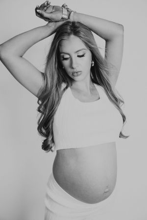 Black and white portrait of a pregnant woman in a white crop top with arms raised, looking down at her baby bump.