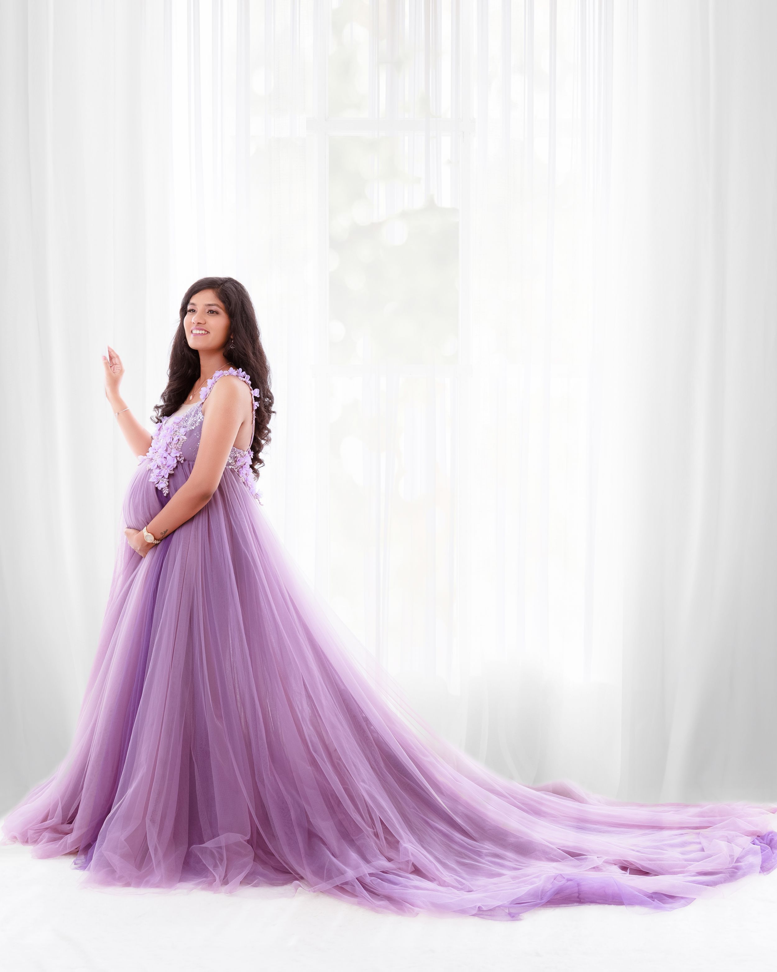 Elegant maternity photoshoot featuring a flowing lavender tulle gown with long train against white curtained backdrop.