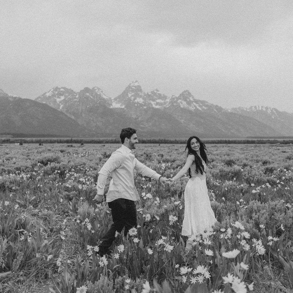 A couple stands in a field of wildflowers with snow-capped Grand Teton mountains in the background.