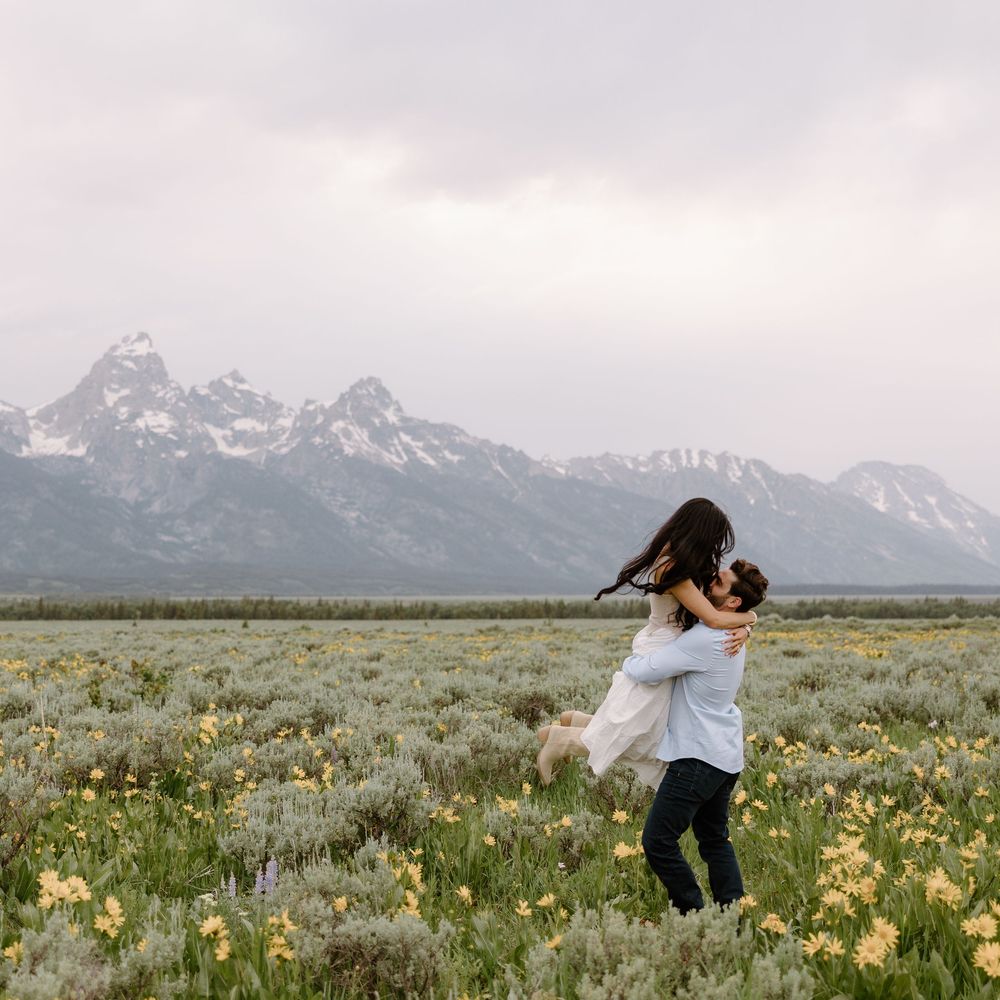 Mountain landscape with yellow wildflowers blooming in meadow beneath snow-capped Grand Teton peaks at sunset.