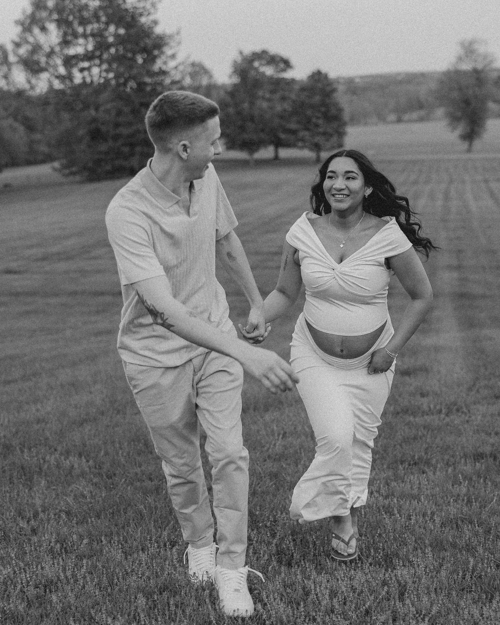 A couple in white outfits playfully runs and laughs together across a grassy field in black and white photography.