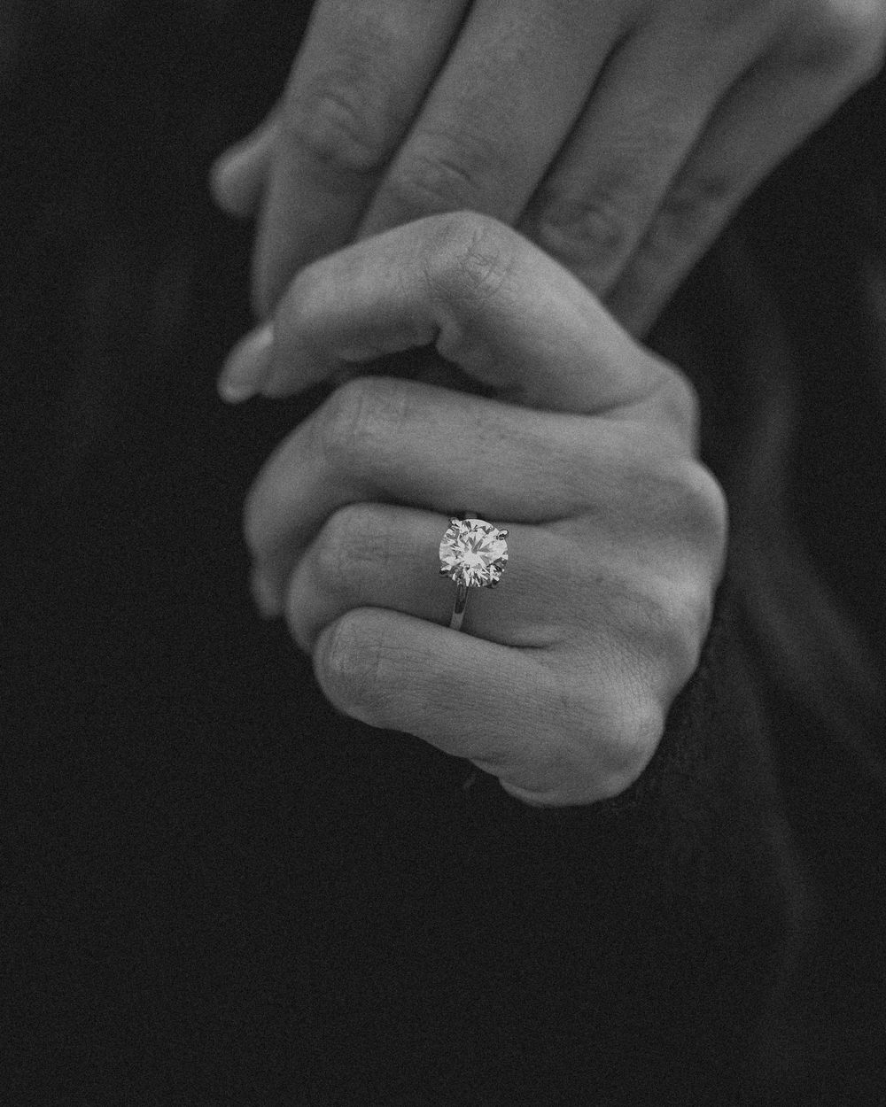 A monochrome image showing hands clasped together with a sparkling diamond engagement ring.