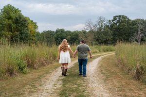 Couple holding hands walking down a rustic dirt path surrounded by tall grass and trees in a scenic outdoor setting.
