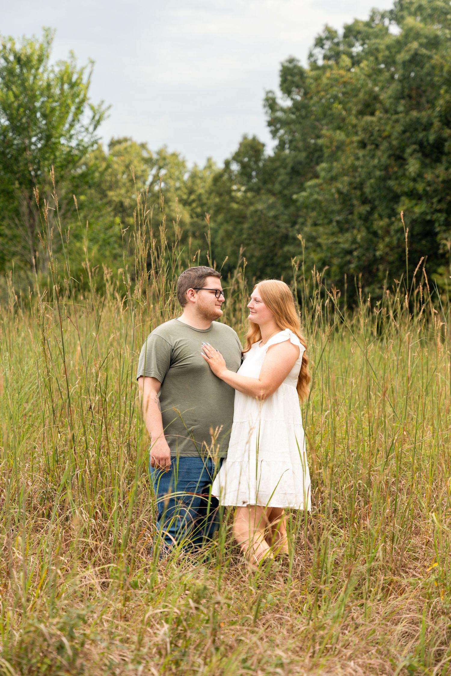 Couple standing in tall grass field, woman in white dress and man in green shirt sharing a romantic moment outdoors.