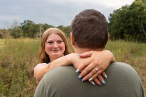 Couple embracing in a field, woman with blue nails smiling over man's shoulder in outdoor engagement photo.