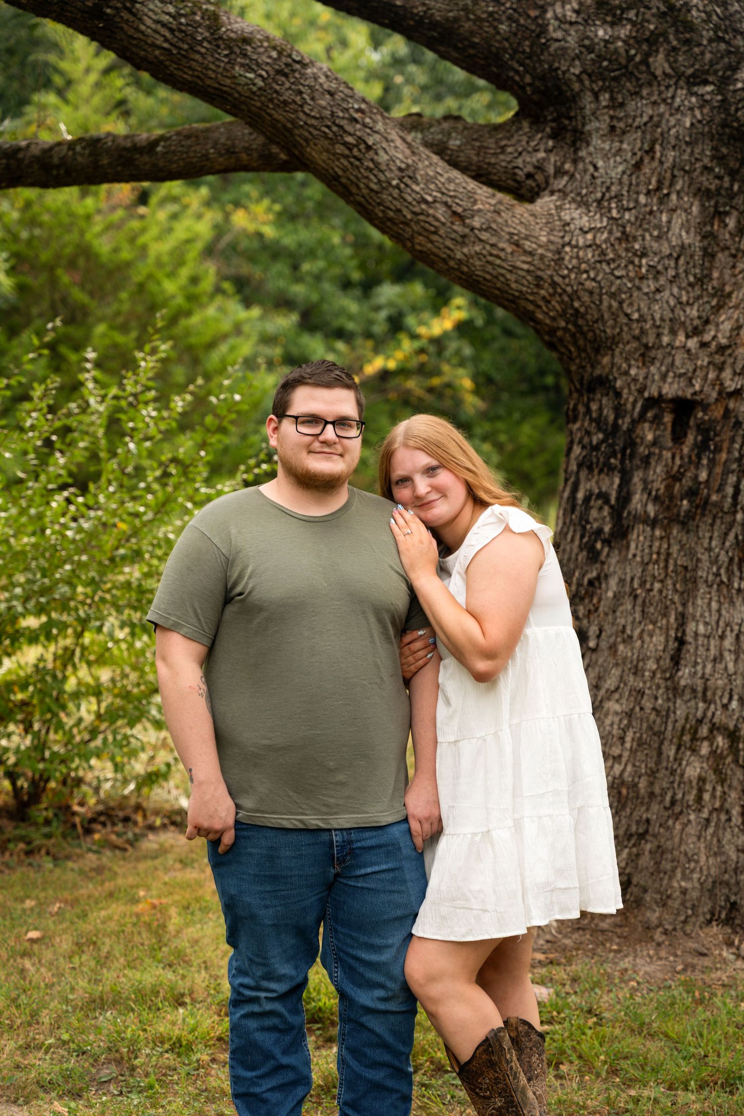 A couple poses outdoors near a large oak tree, man in green shirt and jeans, woman in white dress and cowboy boots.