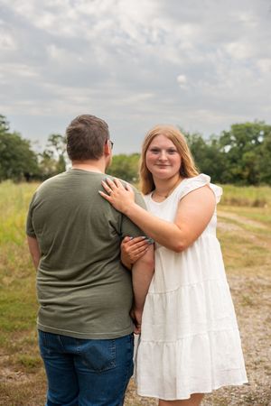 A couple poses outdoors on a gravel path, woman in white dress smiling at camera, man in green shirt facing away.