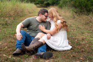 Couple sitting together in a grassy field, sharing a romantic moment, man in green shirt and jeans, woman in white dress.
