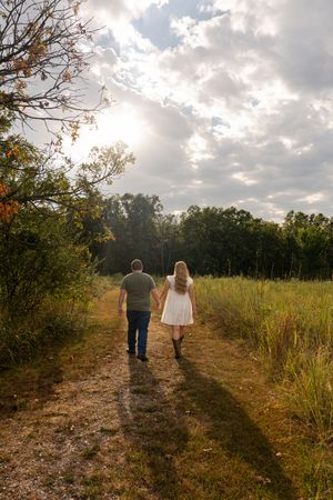 Couple holding hands walking down a rural dirt path surrounded by lush green fields and trees under a cloudy sky.