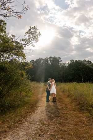 Couple kissing on a sunlit dirt path surrounded by lush green fields and trees on a partly cloudy day.