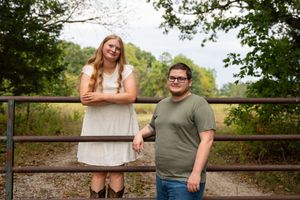 Two young people smiling outdoors near a wooden fence with lush green trees in the background.