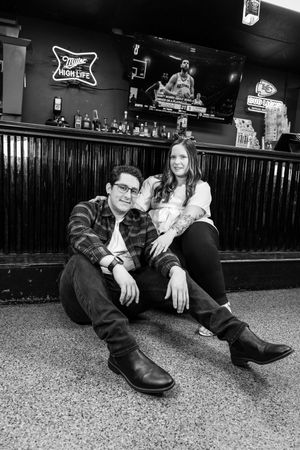Black and white photo of a couple sitting on a bar floor, with Miller High Life neon sign and TV in background.