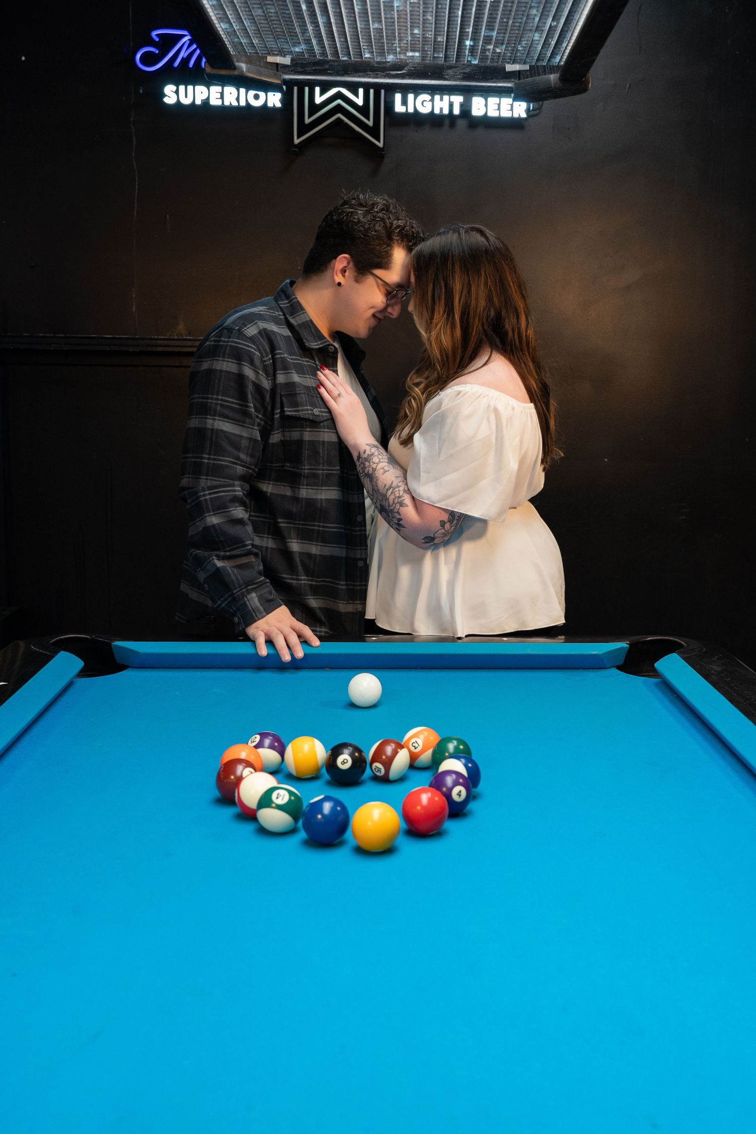 Couple sharing a romantic moment over a blue pool table with billiard balls arranged in a heart shape at a bar.