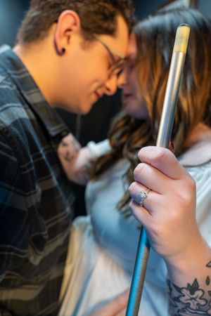 Couple sharing a romantic moment, woman displaying engagement ring while holding a pool cue, tattoos visible.