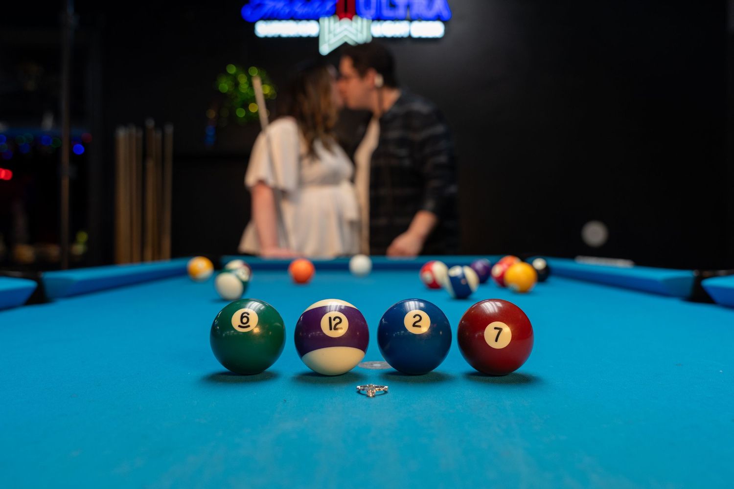 Couple kissing behind pool table with colorful billiard balls in foreground on blue felt surface.