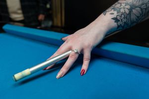 Tattooed hand with red nails resting on a blue pool table beside a billiard cue stick.