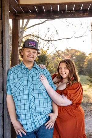 Smiling couple posing under rustic wooden structure, man in turquoise plaid shirt and woman in rust dress.