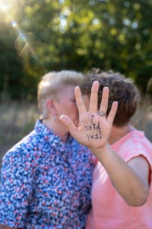 Same-sex couple kissing outdoors, one partner showing hand with engagement ring and 'I said yes' written on palm.