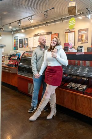 A smiling couple poses together inside a cozy coffee shop, woman in red skirt and boots, man in gray sweater.