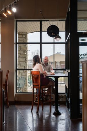 A man and woman sit at a high table by large windows in a modern cafe, engaged in conversation.