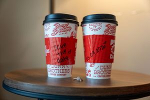 Two Dunn Brothers Coffee red and white to-go cups with black lids sit on a wooden table.
