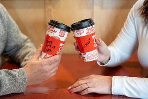 Two people toasting with red Dunkin' Donuts coffee cups at a table, celebrating with hot beverages.