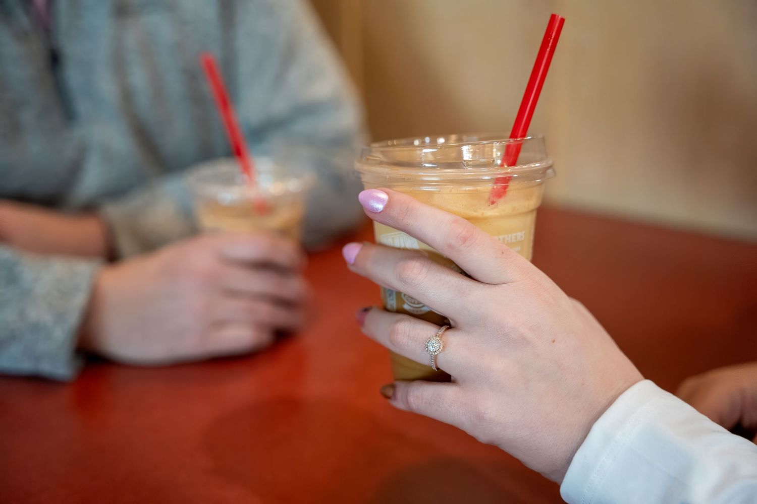 Two people holding iced coffee drinks with red straws in clear plastic cups on a red table.