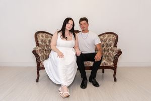 A man and woman sitting together on a vintage floral loveseat, woman in white dress, man in gray shirt and black pants.