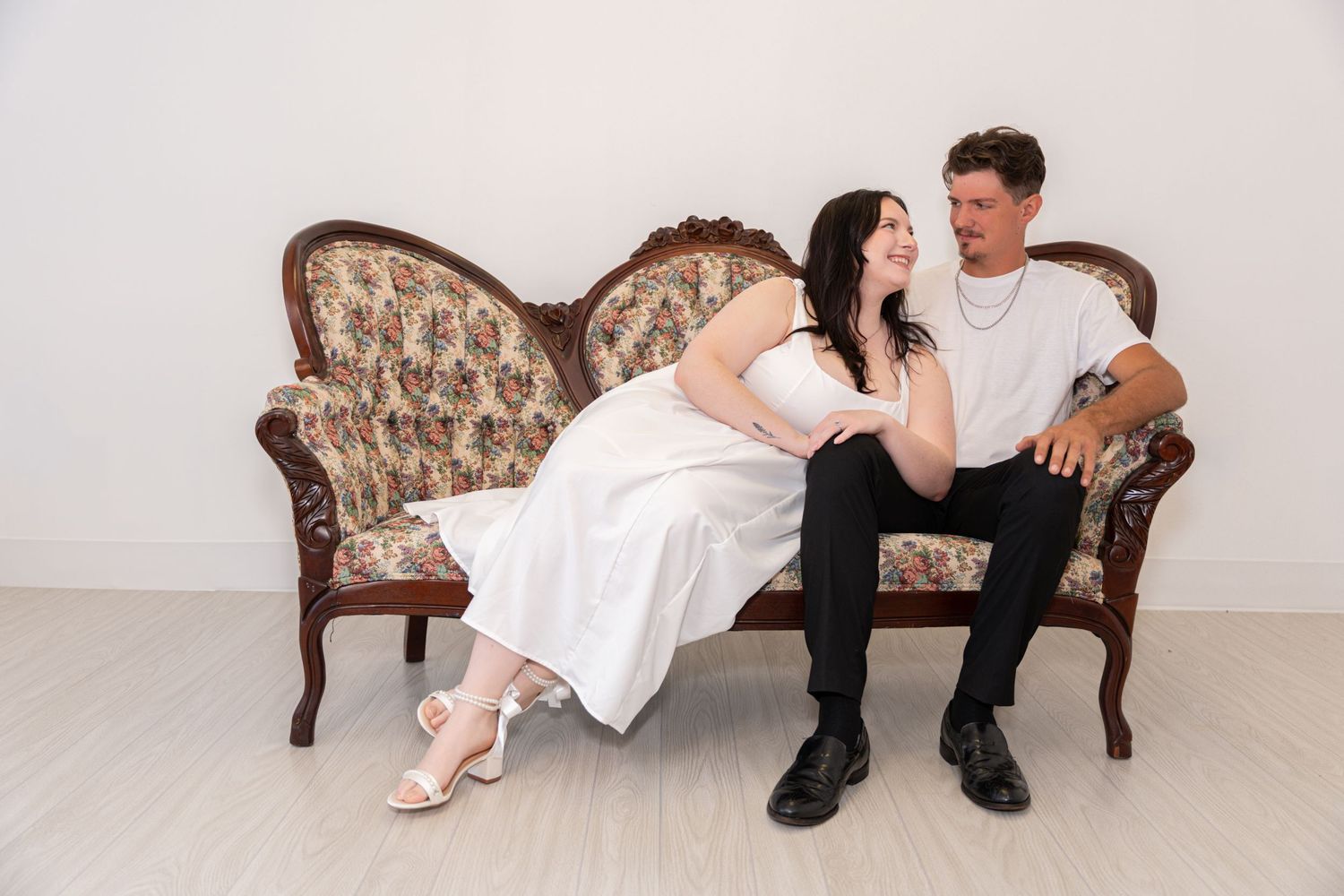 Couple in wedding attire sitting on ornate vintage floral sofa, smiling at each other in bright white studio.
