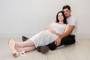 Pregnant couple sitting on floor, woman in white dress leaning against man in white t-shirt, maternity photo session.