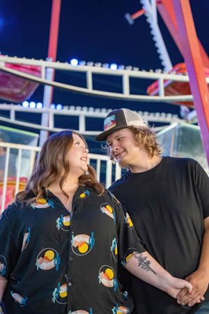 Happy couple holding hands and smiling at each other in front of lit fairground ride at night.