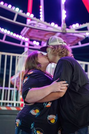 Couple kissing romantically in front of illuminated purple ferris wheel at night carnival.