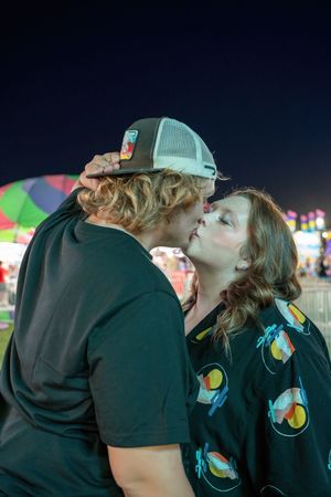 Young couple sharing a kiss at outdoor carnival with colorful night lights in background.