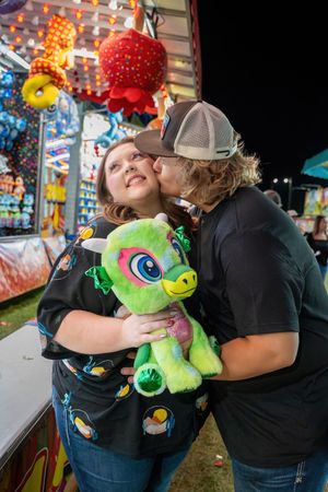 Man kissing woman on cheek as she holds green stuffed animal prize at carnival game booth in Lebanon, Missouri