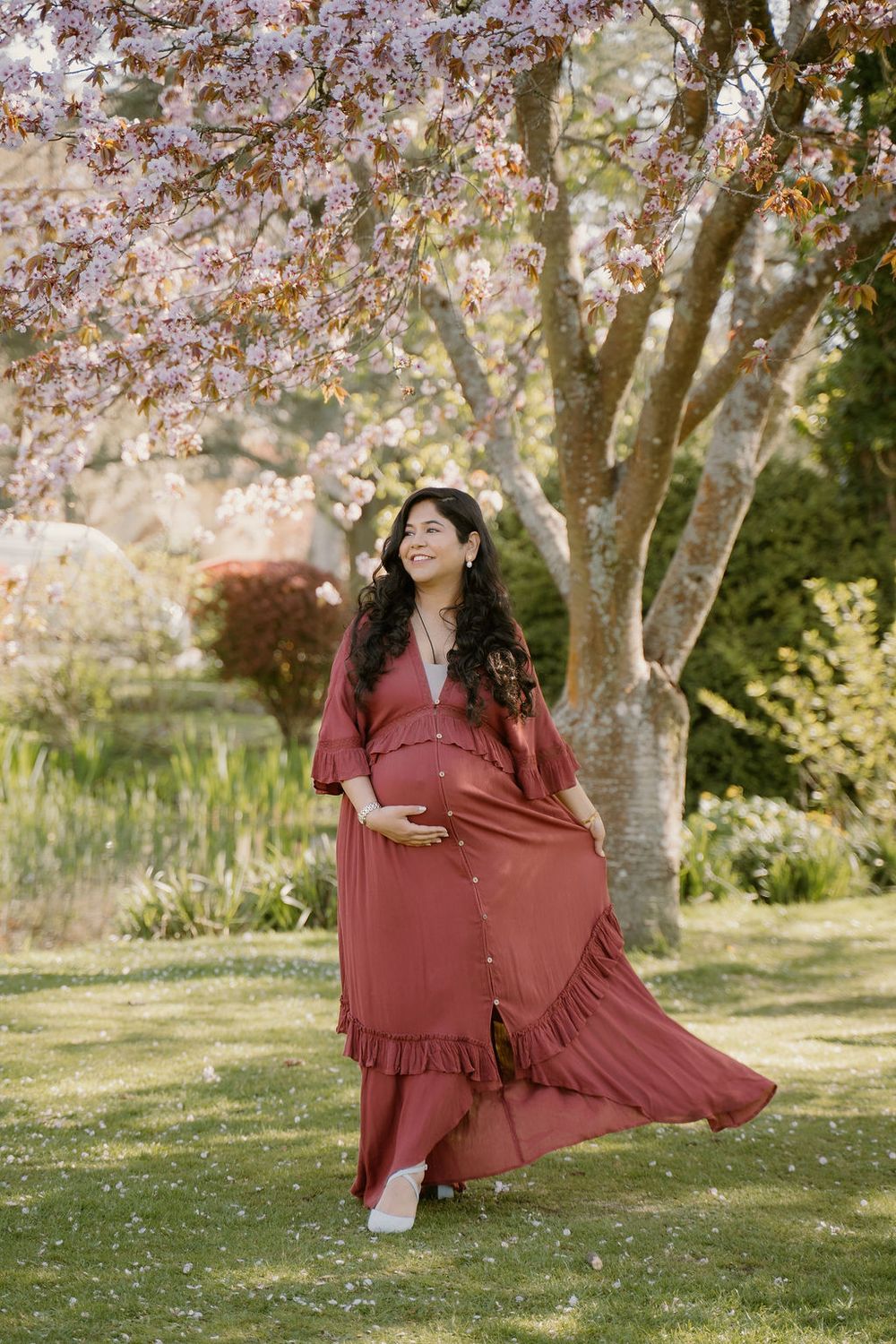 Pregnant mother in a flowing burnt orange dress  holding bump and whooshing her dress in front of a gorgeous cherry blossom tree