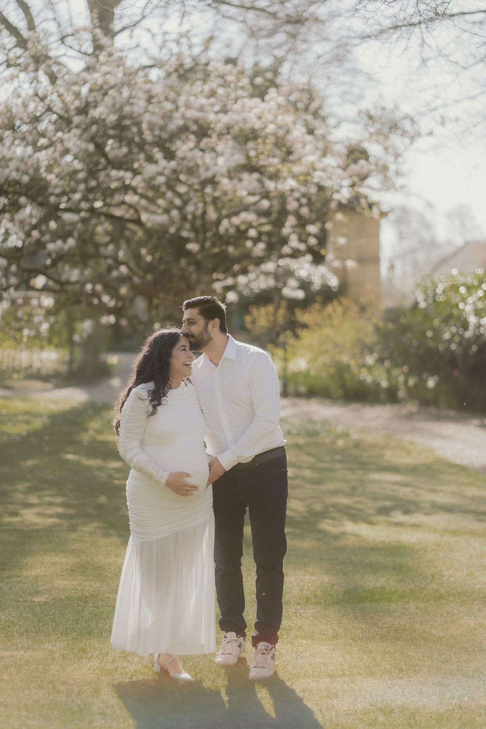 Pregnant mother in a flowing white dress walking cuddling in with her partner, holding bump in front of a gorgeous blossom tree