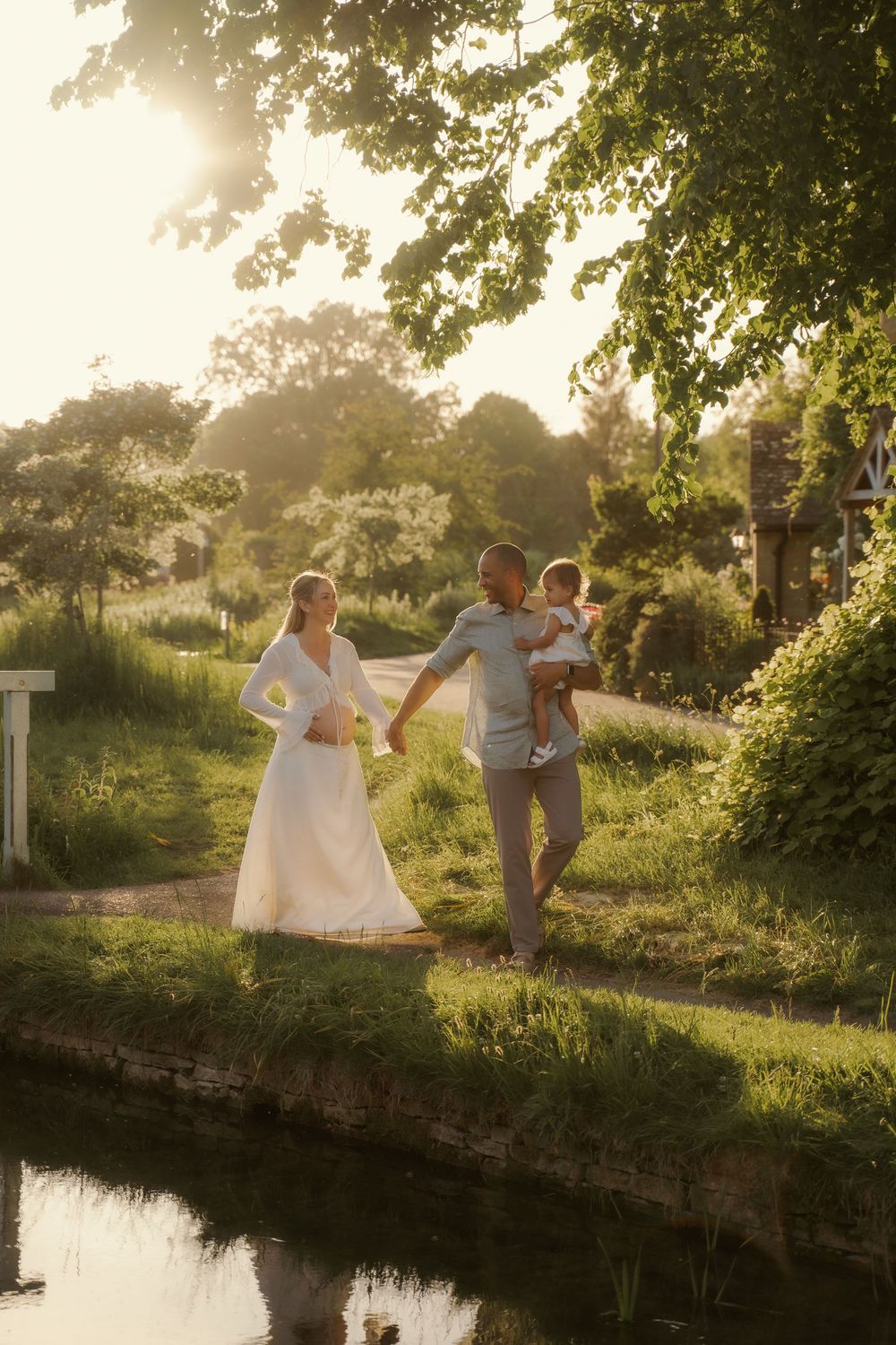Family maternity photoshoot featuring expectant parents and their toddler walking beside a tranquil stream in warm golden evening light.