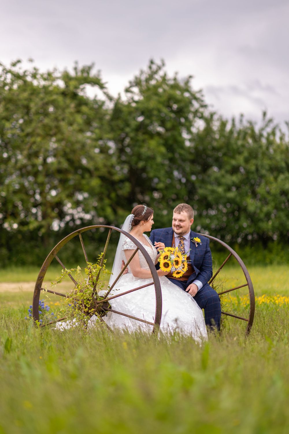 A couple sitting by an antique wagon wheel with sunflowers in a rustic field setting.