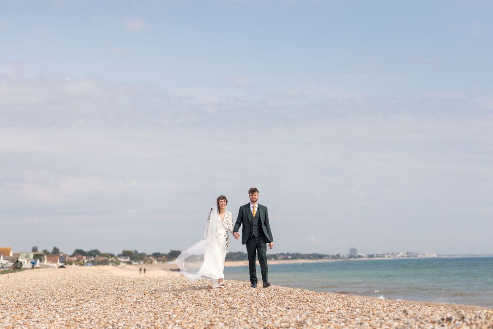 A couple walking along a seaside beach with waves and coastal buildings in the background.