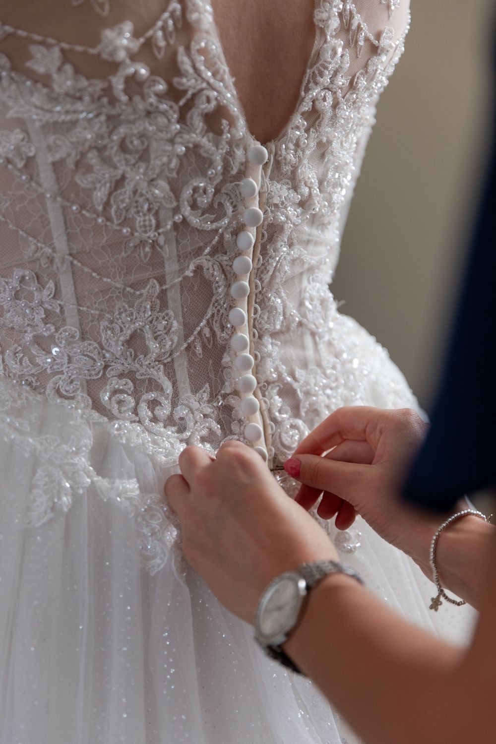 Close-up of delicate lace buttons being fastened on the back of an ornate white wedding dress.