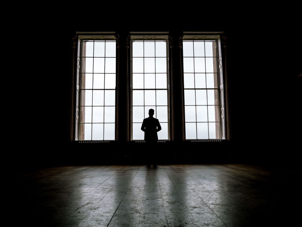 Silhouetted figure stands centered between three large windows in a dark room.