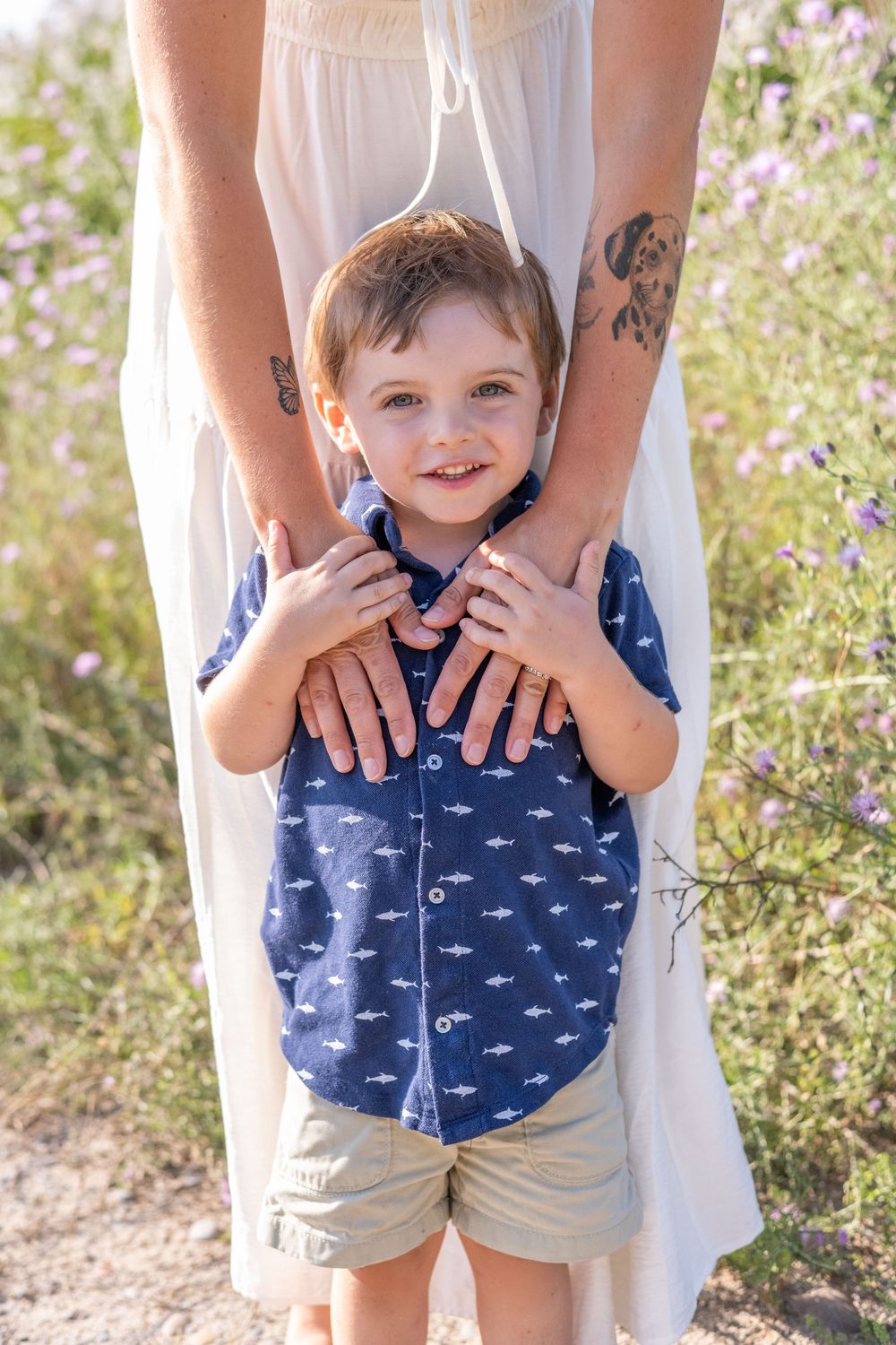 A child wearing a blue patterned shirt and khaki shorts stands against a backdrop of wildflowers.