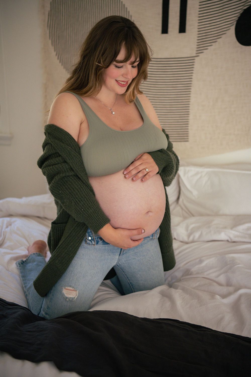 A pregnant woman in a green cardigan and jeans relaxes on white bedding.